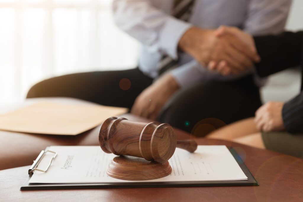 A wooden gavel rests on a contract on a table, symbolizing a legal agreement, as two people shake hands in the background.