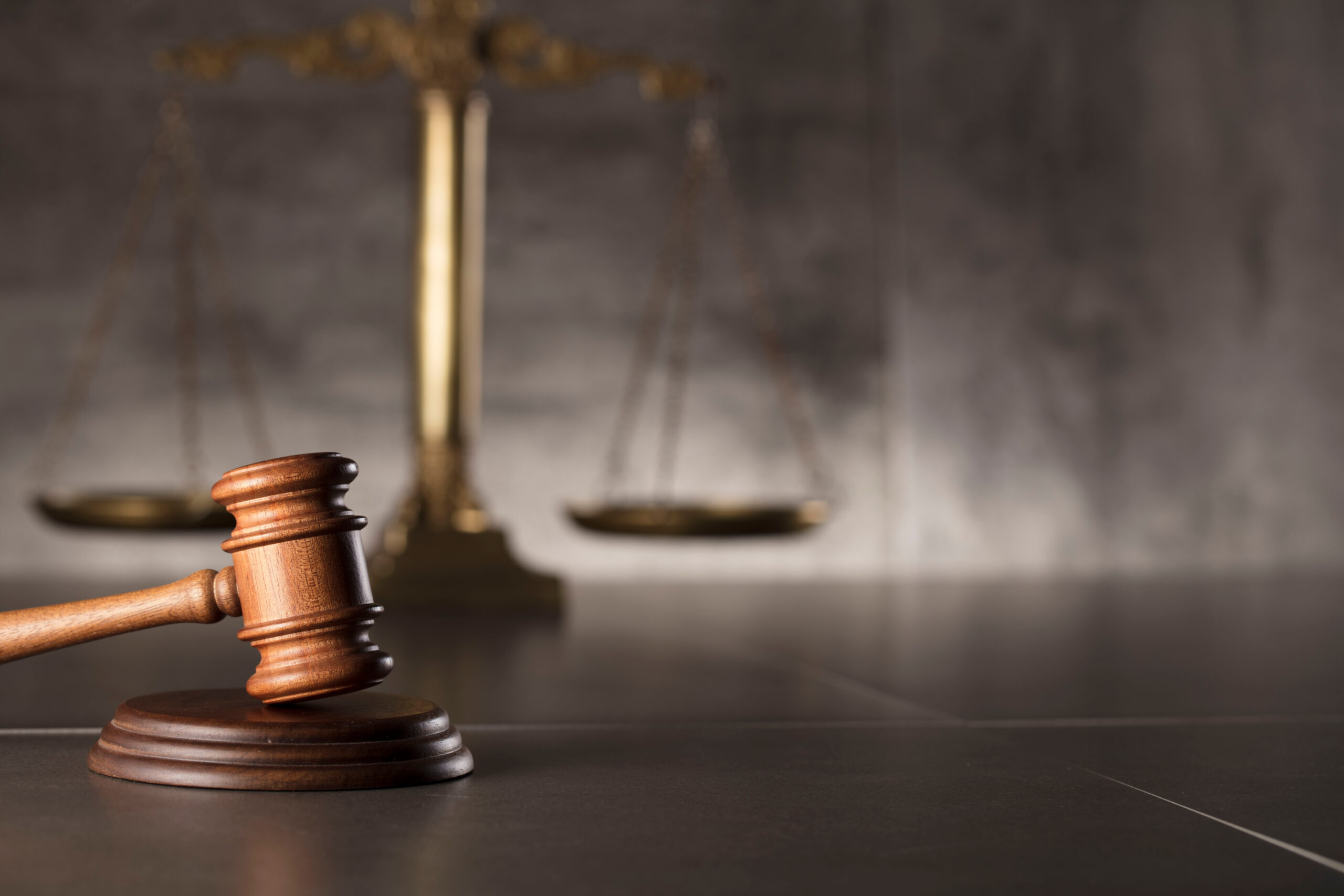 A wooden gavel rests on a dark surface, with a blurred golden balance scale in the background, symbolizing justice and the legal system.